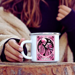 K&ouml;nnte beinhalten: Wei&szlig;er Emaillebecher mit silbernem Rand. Er zeigt eine rosa-schwarze Illustration von zwei sich umarmenden Skeletten, umrahmt von Blumen und Sternen. Die Texte "AMOR" und "THE LOVERS" sind ebenfalls sichtbar.