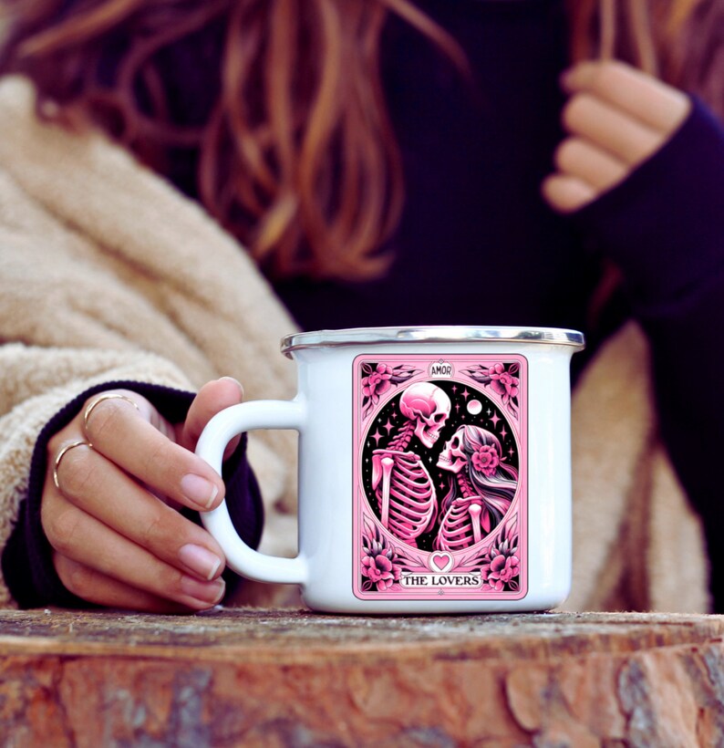 K&ouml;nnte beinhalten: Wei&szlig;er Emaillebecher mit silbernem Rand. Er zeigt eine rosa-schwarze Illustration von zwei sich umarmenden Skeletten, umrahmt von Blumen und Sternen. Die Texte "AMOR" und "THE LOVERS" sind ebenfalls sichtbar.