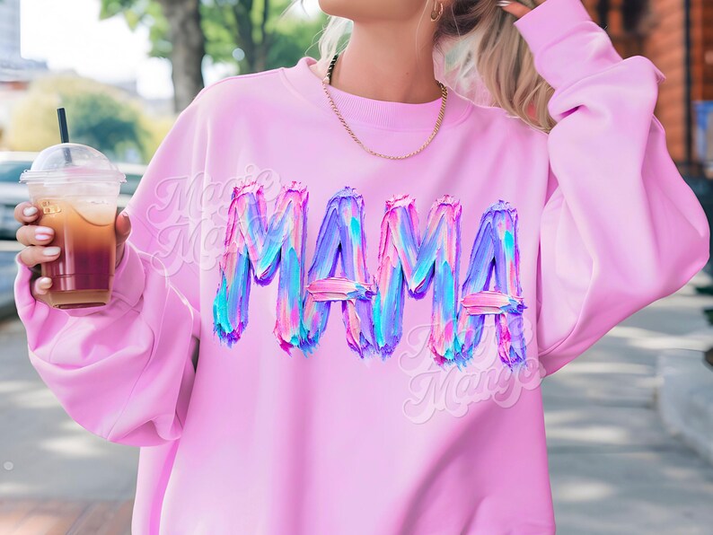 May include: A pale pink sweatshirt featuring the word "MAMA" in a vibrant, textured design. The letters are rendered in shades of blue, purple, and pink. The sweatshirt has long sleeves and a round neck.