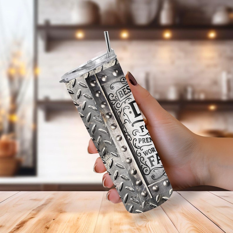 May include: A stainless steel tumbler with a diamond plate design and the text "BEST DAD EVER". The tumbler has a silver straw and a clear lid. The tumbler is held in a hand.
