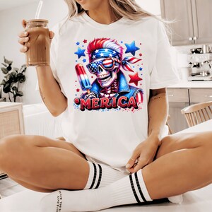 May include: White t-shirt with a graphic of a skeleton wearing a bandana and sunglasses, holding a red, white, and blue popsicle. The graphic is surrounded by stars and the word "MERICA" is printed below the skeleton.