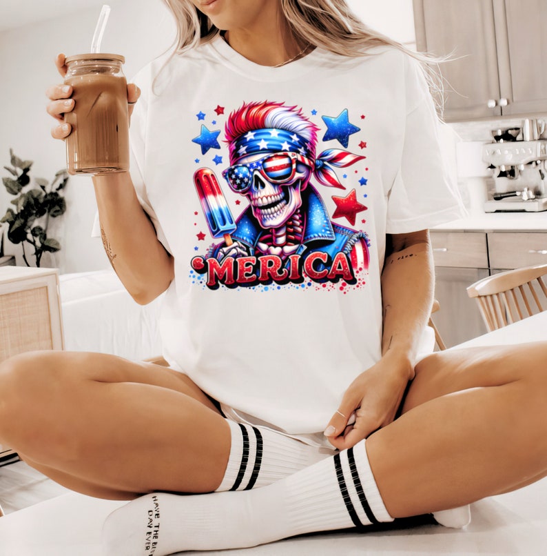 May include: White t-shirt with a graphic of a skeleton wearing a bandana and sunglasses, holding a red, white, and blue popsicle. The graphic is surrounded by stars and the word "MERICA" is printed below the skeleton.