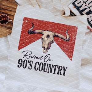 May include: White tote bag with a red and white graphic of a bull skull with the text "Raised On 90's Country".