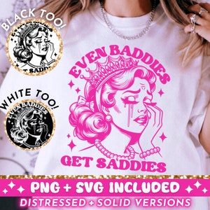May include: A white t-shirt with a pink graphic design featuring a woman with a crown and the text "Even Baddies Get Saddies". The graphic is surrounded by stars and glitter. The text "Black Too!" and "White Too!" are in circles at the top of the image.