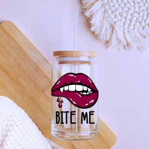 May include: A clear glass reusable drinking cup with a bamboo lid and a straw. The cup has a graphic of a red vampire mouth with fangs and dripping blood. The text "Bite Me" is printed below the graphic.