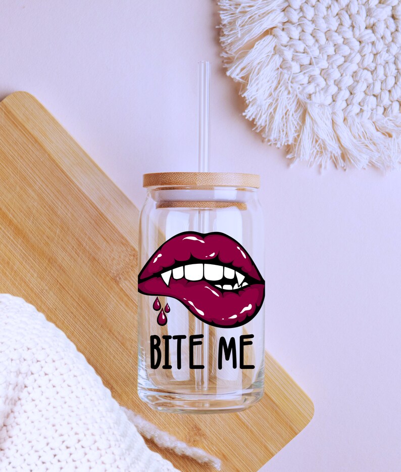 May include: A clear glass reusable drinking cup with a bamboo lid and a straw. The cup has a graphic of a red vampire mouth with fangs and dripping blood. The text "Bite Me" is printed below the graphic.