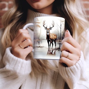 May include: White ceramic mug with a brown and white deer illustration. The deer is standing in a lake with trees in the background.