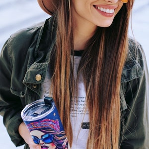 May include: A stainless steel tumbler with a colorful, psychedelic design featuring a woman wearing sunglasses and a headscarf. The tumbler has a lid with a straw.