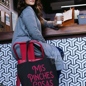 May include: A black tote bag with the text "Mis Pinches Cosas" in pink lettering. The bag is sitting on a red chair in front of a black and white geometric pattern.