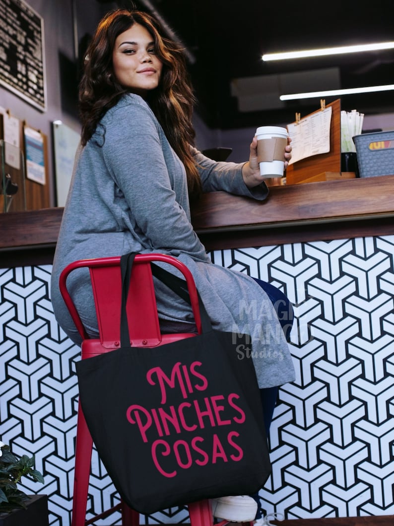 May include: A black tote bag with the text "Mis Pinches Cosas" in pink lettering. The bag is sitting on a red chair in front of a black and white geometric pattern.