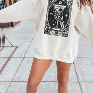 May include: A white sweatshirt with a black and white illustration of a skeleton hand holding a martini glass. The illustration is framed in a black border with the text "THE MARTINI" at the bottom.
