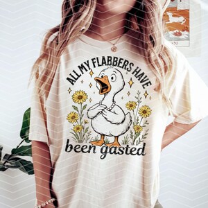 以下が含まれることがあります： 漫画の鴨と花が描かれた白いTシャツ。シャツのテキストは「All my flabbers have been gasted」です。
