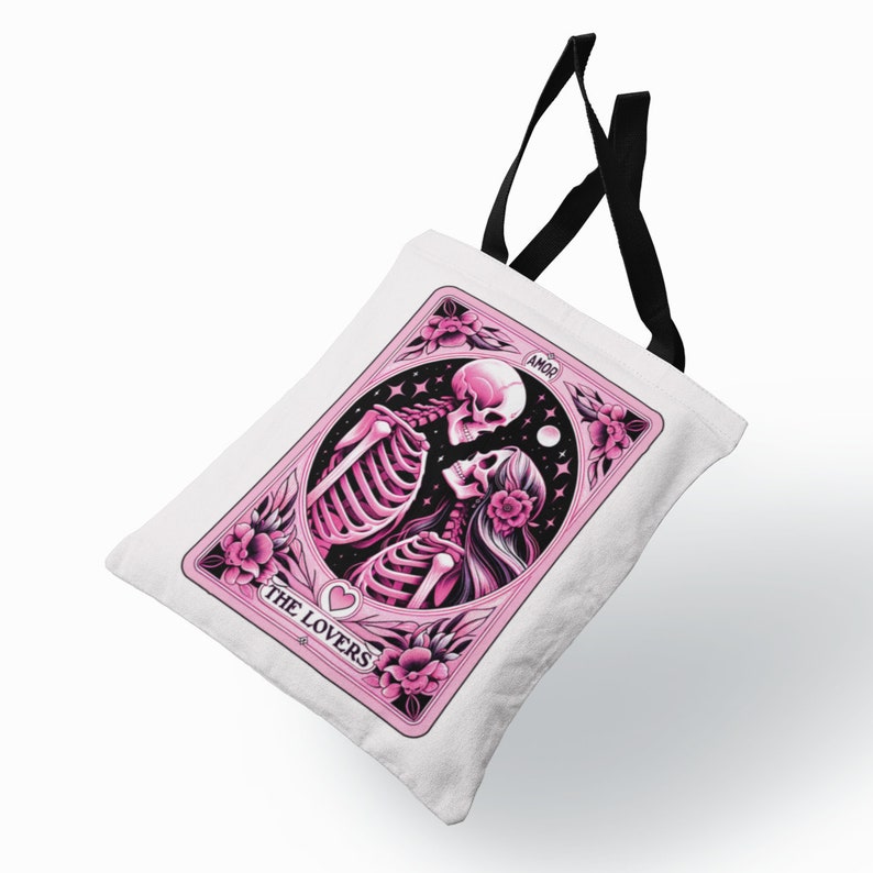 K&ouml;nnte beinhalten: Wei&szlig;e Tragetasche mit einem rosa Tarotkarten-Design. Das Design zeigt Skelette, Blumen und die Worte "THE LOVERS" und "AMOR". Sie hat schwarze Griffe.