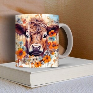 May include: A white ceramic mug with a brown and white cow's face surrounded by orange, white, and blue flowers. The mug has a white handle.