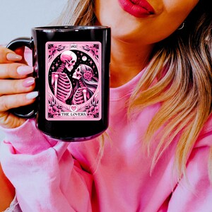 K&ouml;nnte beinhalten: Schwarze Keramik-Tasse mit einem rosa-schwarzen Tarotkarten-Design. Die Karte zeigt Skelette, Blumen und die Worte "THE LOVERS" und "AMOR". Die Tasse hat einen gro&szlig;en Henkel und ist vor einem rosa Hintergrund.