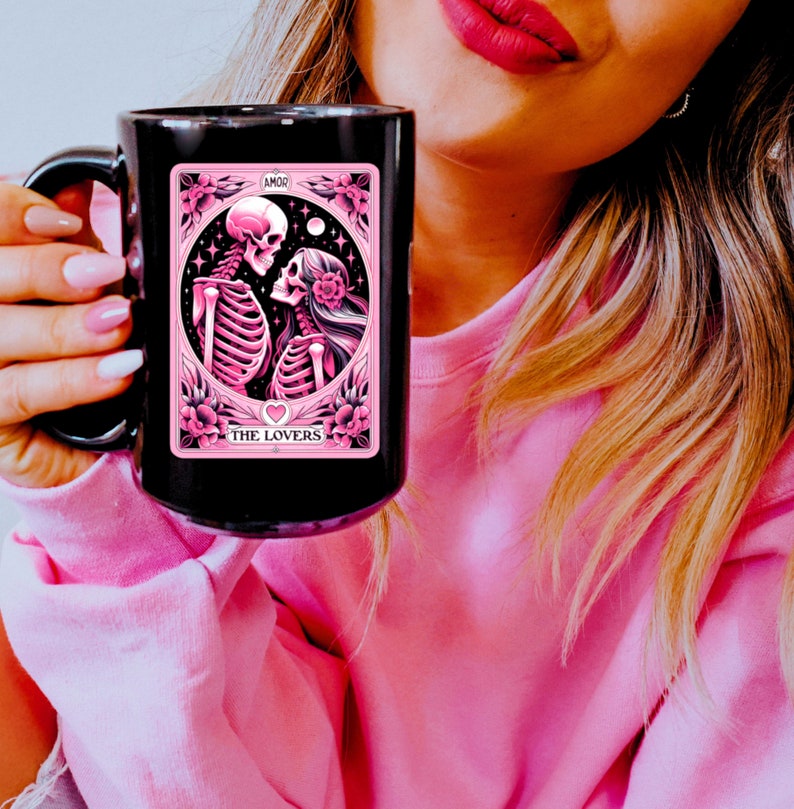 K&ouml;nnte beinhalten: Schwarze Keramik-Tasse mit einem rosa-schwarzen Tarotkarten-Design. Die Karte zeigt Skelette, Blumen und die Worte "THE LOVERS" und "AMOR". Die Tasse hat einen gro&szlig;en Henkel und ist vor einem rosa Hintergrund.