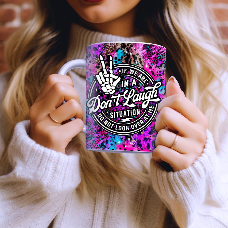 May include: A white ceramic mug with a pink, blue, and black design. The mug features a peace sign with a skeleton hand, and the text "If we are in a situation Don't Laugh Do not look over at me".