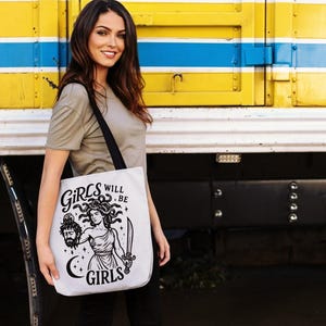 May include: White tote bag with a black strap featuring a black and white illustration of Medusa holding a sword and a severed head. The text "GIRLS WILL BE GIRLS" is printed on the bag in a stylized font. The bag is held by a person.