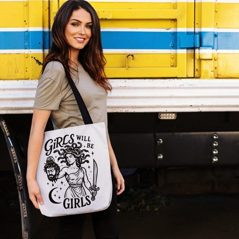 May include: White tote bag with a black strap featuring a black and white illustration of Medusa holding a sword and a severed head. The text "GIRLS WILL BE GIRLS" is printed on the bag in a stylized font. The bag is held by a person.