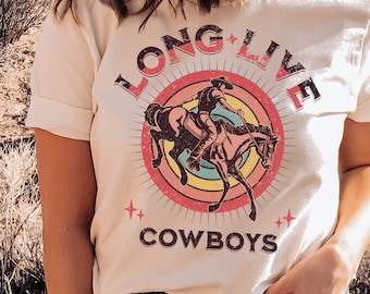 Puede incluir: Una camiseta blanca con un gráfico de estilo vintage de un vaquero montando un caballo. El gráfico está rodeado de un círculo de colores arcoíris con el texto "Long Live Cowboys" en fuente negra.