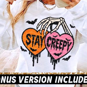 May include: White sweatshirt with a Halloween design. The design features two heart-shaped graphics with the words "STAY CREEPY" in black, held by a skeleton hand, with bat silhouettes and dripping details.