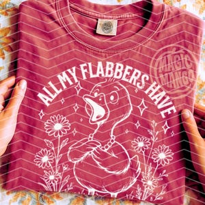 以下が含まれることがあります： ピンクのTシャツに、白いダックのグラフィックがプリントされています。ダックの周りに「All my flabbers have」というテキストが書かれています。グラフィックには、花と星も含まれています。
