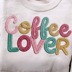 May include: A light beige sweatshirt with the embroidered text "Coffee Lover" in a colorful, textured font. The letters are in shades of pink, yellow, and teal, creating a visually appealing design.