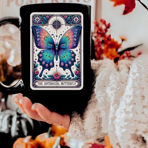 May include: Black coffee mug with a white design of a butterfly with iridescent wings and the text "The Antisocial Butterfly".