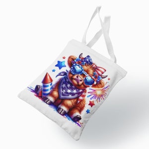 May include: A white tote bag with a cartoon Highland cow wearing red, white, and blue sunglasses and a bandana. The cow is holding a red, white, and blue firework. There are stars and fireworks in the background.