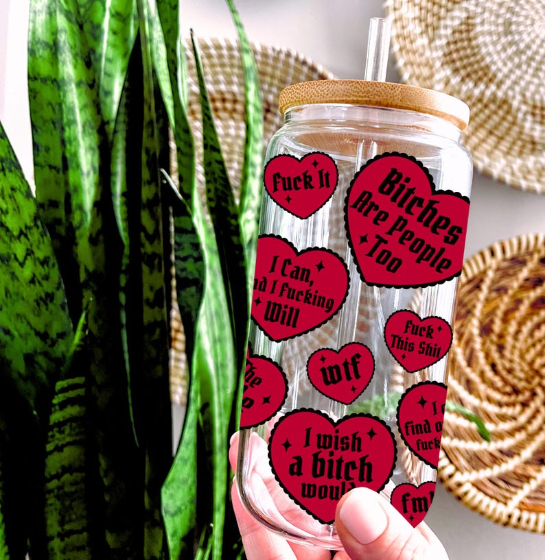 Puede incluir: Un vaso de vidrio transparente con tapa y pajita de bamb&uacute;, adornado con pegatinas rojas en forma de coraz&oacute;n. Las pegatinas presentan varias frases en texto blanco, incluyendo "Fuck It" y "Bitches Are People Too."