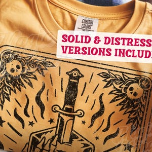 Pu&ograve; includere: T-shirt giallo senape con grafica nera invecchiata raffigurante un pugnale, teschi ed elementi floreali. La maglietta include il testo "SOLID & DISTRESSED VERSIONS INCLUDED" su un'etichetta bianca.
