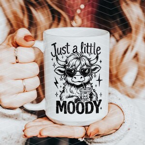 May include: White ceramic mug with a black and white illustration of a Highland cow wearing sunglasses and drinking from a cup. The text "Just a little MOODY" is printed on the mug.
