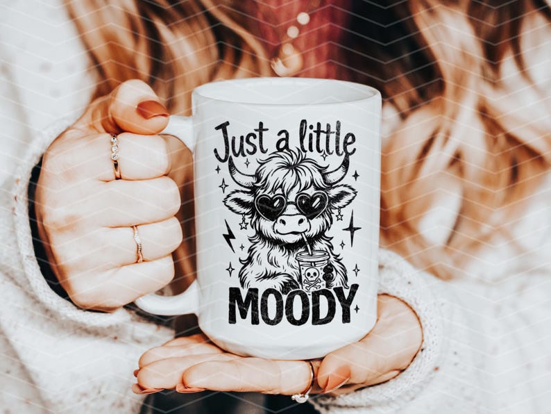 May include: White ceramic mug with a black and white illustration of a Highland cow wearing sunglasses and drinking from a cup. The text "Just a little MOODY" is printed on the mug.