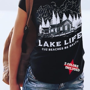 May include: Black t-shirt with a white graphic of a lakeside cabin, trees, and mountains. The text "LAKE LIFE cuz beaches be salty" is printed below the graphic. A red starburst graphic indicates "3 COLORS INCLUDED".