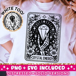 Puede incluir: Camiseta blanca con un diseño gráfico en blanco y negro de una carta de tarot con un diamante y una mano de esqueleto. El texto "Crystal Energy" está en la carta. El texto "White Too!" está en un círculo dorado en la camiseta.
