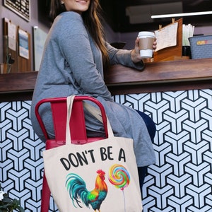 May include: A white canvas tote bag with the text "Don't be a" and a colorful rooster illustration holding a lollipop.