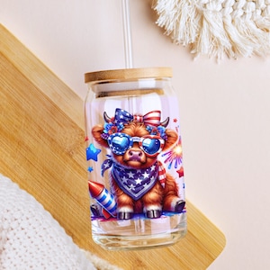 May include: A glass tumbler with a bamboo lid and a straw. The tumbler features a cartoon illustration of a brown cow wearing sunglasses and a bandana with stars and stripes. The cow is surrounded by stars, fireworks, and a rocket.
