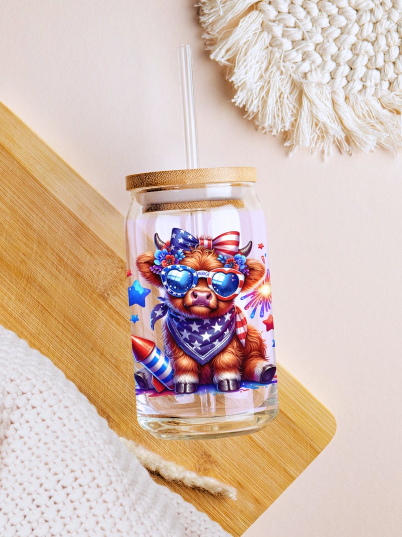 May include: A glass tumbler with a bamboo lid and a straw. The tumbler features a cartoon illustration of a brown cow wearing sunglasses and a bandana with stars and stripes. The cow is surrounded by stars, fireworks, and a rocket.