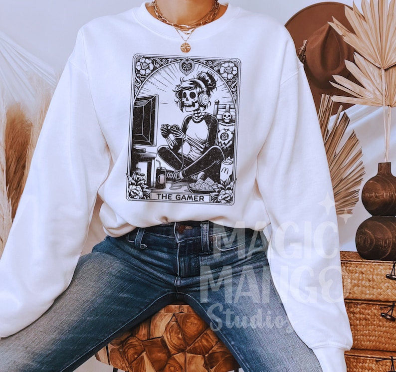 Op de afbeelding: Witte trui met een zwart-witte illustratie van een skelet dat videogames speelt. De illustratie is omlijst met een decoratieve rand en bevat de tekst "The Gamer".