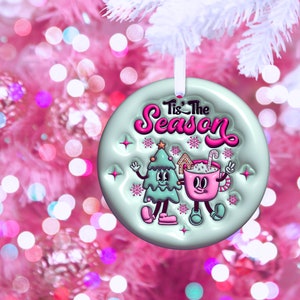 Puede incluir: Un adorno navide&ntilde;o de color azul claro y verde con un &aacute;rbol de dibujos animados y una taza de chocolate caliente. El adorno tiene el texto "Tis' the Season" en letras rosas.