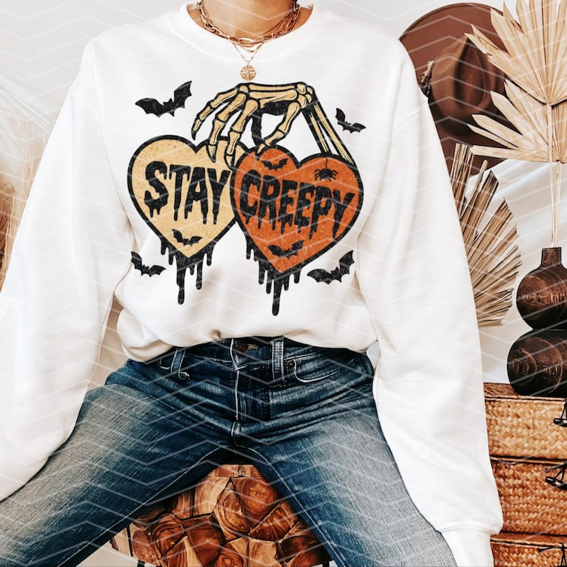 May include: White sweatshirt with a Halloween graphic. The design features two heart-shaped pumpkins with the words "STAY CREEPY" and a skeleton hand holding them. Black bats are scattered around the hearts.