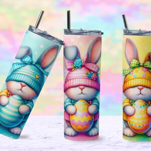 May include: Three Easter-themed stainless steel tumblers with a pastel background. Each tumbler features a cartoon bunny wearing a hat and holding an Easter egg. The bunny on the left is wearing a blue hat, the bunny in the middle is wearing a pink hat, and the bunny on the right is wearing a yellow hat.
