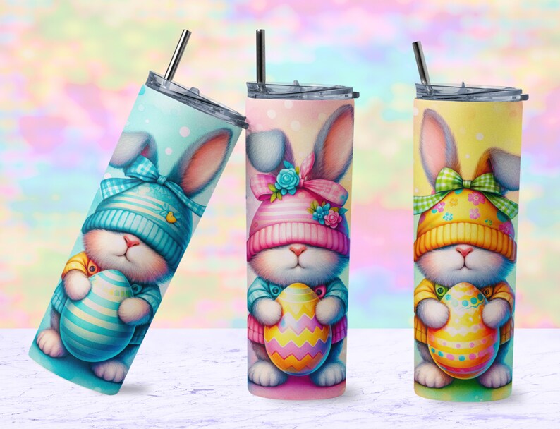 May include: Three Easter-themed stainless steel tumblers with a pastel background. Each tumbler features a cartoon bunny wearing a hat and holding an Easter egg. The bunny on the left is wearing a blue hat, the bunny in the middle is wearing a pink hat, and the bunny on the right is wearing a yellow hat.