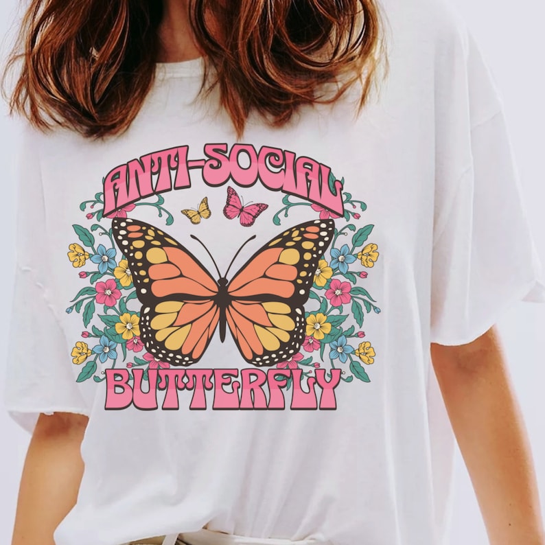Peut inclure: T-shirt blanc avec un motif de papillon rose et orange et le texte "Anti-Social Butterfly" en lettres roses.