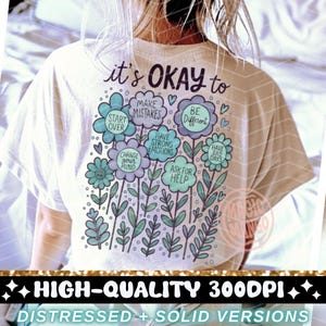 以下が含まれることがあります： 白いTシャツに、花柄のグラフィックデザインと「it's OKAY to」というテキストがプリントされています。テキストには、「make mistakes」、「start over」、「be different」、「have strong emotions」、「change your mind」、「have bad days」、「ask for help」などのフレーズが並んでいます。