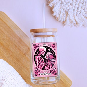 K&ouml;nnte beinhalten: Ein klares Glas mit einem Bambusdeckel und einem Glasstrohhalm. Der Becher zeigt eine rosa und schwarze Grafik von zwei Skeletten mit den Worten "THE LOVERS" und "AMOR" auf dem Design. Der Becher steht auf einer Holzoberfl&auml;che.
