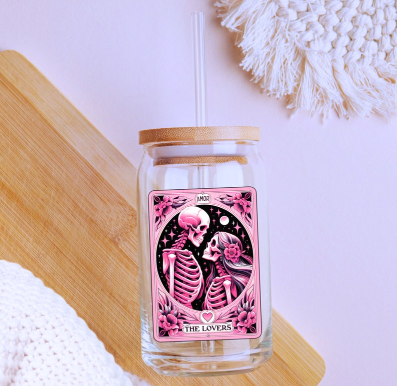 K&ouml;nnte beinhalten: Ein klares Glas mit einem Bambusdeckel und einem Glasstrohhalm. Der Becher zeigt eine rosa und schwarze Grafik von zwei Skeletten mit den Worten "THE LOVERS" und "AMOR" auf dem Design. Der Becher steht auf einer Holzoberfl&auml;che.