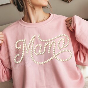 May include: A light pink sweatshirt with the word "Mama" spelled out in faux pearl beads. The sweatshirt has long sleeves and a crew neck. The design is centered on the chest.