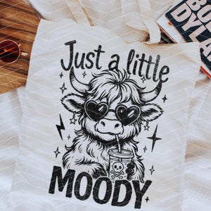May include: A white tote bag with black text and a black and white illustration of a Highland cow wearing heart-shaped sunglasses and drinking from a cup. The text reads "Just a little moody".