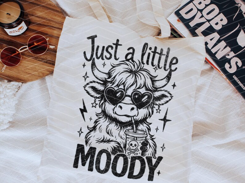 May include: A white tote bag with black text and a black and white illustration of a Highland cow wearing heart-shaped sunglasses and drinking from a cup. The text reads "Just a little moody".